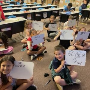 Group of primary school students seated on the classroom floor, each holding a whiteboard with drawings and numbers.