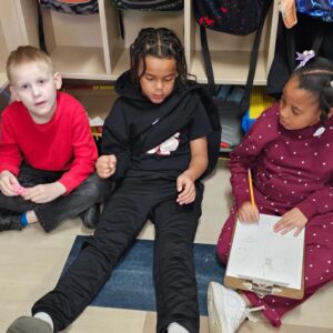 Three children sit on a classroom floor by cubbies; the boy in red holds a small pink object while the girl in maroon-dotted clothing draws on a clipboard with a pencil beside him?