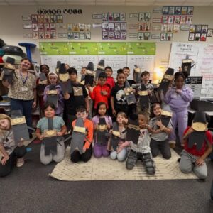Group of diverse elementary students and their teacher posing in a classroom while proudly displaying black paper cutout figures with beige faces they made for a craft project.