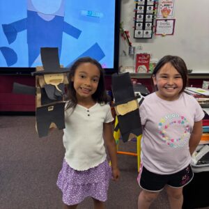 Two smiling girls in a classroom proudly display their homemade cardboard figure crafts with tall black hats.