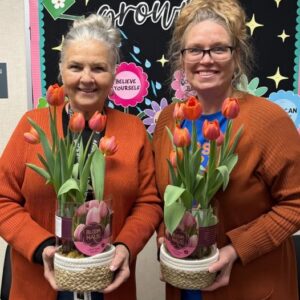 Two smiling women in orange cardigans hold potted orange tulips in woven baskets in front of a colorful bulletin-board backdrop.