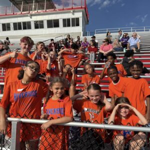 Children in orange Riverside shirts smile and pose on red stadium bleachers, with a chain-link fence in the foreground and spectators behind them.